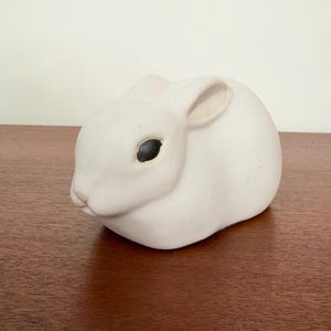 May include: A small, matte white ceramic figurine of a rabbit in a resting position. The rabbit has a smooth, rounded body, pointed ears folded back, and a single visible dark, oval-shaped eye. The surface of the figurine is plain and unadorned.
