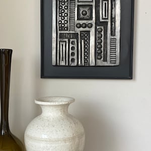 May include: A black and silver geometric wall sculpture with raised circles and lines. The sculpture is framed in black and is hanging on a white wall.