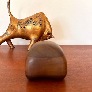 May include: A gold-colored bull figurine with a distressed black finish and a smooth, dark brown wooden box with a rounded top. The bull is positioned behind the box, showing its tail raised and its head lowered in a charging stance.