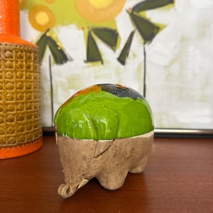 May include: A small, handmade ceramic sculpture with a rounded, green top and a textured, brown base. The sculpture has a unique, organic shape and is displayed on a wooden surface. The background includes a colorful abstract painting and a decorative orange vase.