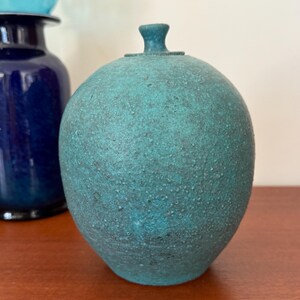 May include: A teal-colored, textured ceramic jar with a lid. The jar has a rounded shape and a small, cylindrical handle on top. A dark blue vase is partially visible in the background.