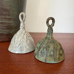 May include: Two ceramic bells with loop handles sit on a wooden surface. One bell is light gray with a textured pattern, and the other is green with a mottled appearance. The bells have a conical shape.