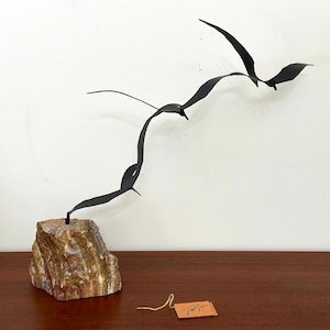 May include: A black metal sculpture of three birds in flight, mounted on a natural stone base. The birds have a streamlined design, with outstretched wings. The stone base has a mix of brown, beige, and gray tones. A small tag hangs from the base.