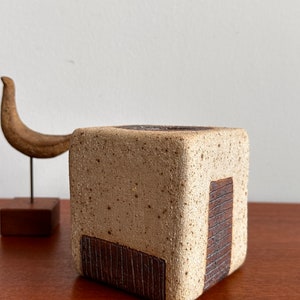 May include: A square, beige ceramic vase with a textured surface and dark brown accents. The vase has a rectangular, ridged design on one side. A wooden sculpture is in the background.