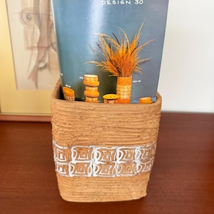 May include: A brown ceramic planter with a white geometric design. The planter holds a blue brochure with the text "Sunstone DESIGN 30" and dried ornamental grass. Decorative ceramic pieces are also visible.