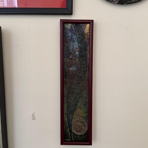 May include: A tall, narrow rectangular wall art piece framed in dark red wood. The artwork features a textured, dark, curved vertical shape set against a mottled background of deep blue, brown, and green tones, with a small, circular, light-colored textured element near the bottom.