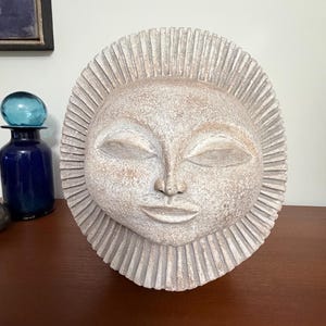 May include: A circular stone-like sculpture featuring a stylized sun face with closed eyes, a small nose, and a serene mouth. The face is surrounded by a radiating border of vertical ridges, finished in a textured, light beige and tan matte color.