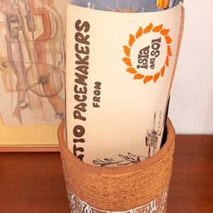 May include: A brown ceramic pot with a white and brown patterned band holds a brochure with the text "Isla del Sol" and "Atio Pacemakers". The brochure has an orange sun logo. A framed artwork is in the background.