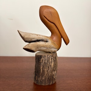 May include: A hand-carved wooden sculpture of a pelican perched on a cylindrical log base. The bird features a smooth, polished light brown head and beak, contrasting with the textured, weathered, and light-colored wood of its body and wings.