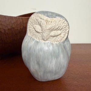 May include: A ceramic owl figurine with a smooth, glossy light gray and white marbled glaze. The owl has a textured, matte off-white face with closed eyes and a pointed beak. The body is rounded and features a soft, blended color pattern.