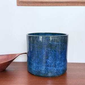 May include: A cylindrical, glazed ceramic planter with a deep blue color gradient. The planter has a textured surface and a wide opening. A wooden bowl is partially visible on the left side, and a painting hangs in the background.
