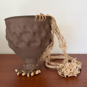 May include: A brown ceramic planter with a textured surface and a decorative macrame plant hanger. The planter has a unique, sculpted design and a small, decorative base. The macrame hanger is beige.