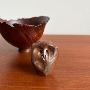 May include: A small, brown ceramic owl figurine with detailed carving. The owl is sitting on a wooden surface.