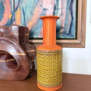 May include: A vintage orange and yellow ceramic vase. The vase has a cylindrical body with a textured, repeating pattern in yellow and a smooth orange neck. The vase is displayed on a wooden surface, with a wooden sculpture and a framed painting in the background.