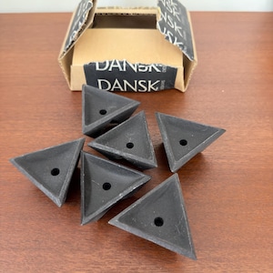 May include: Six matte black triangular candle holders, each featuring a single circular hole in the center. The items are arranged in front of an open cardboard box labeled with the text DANSK DESIGNS on the front and along the top flaps.