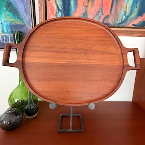 Può includere: Un vassoio rotondo in legno marrone caldo con manici integrati. Il vassoio è sostenuto da un supporto in metallo scuro. Sullo sfondo sono visibili bottiglie di vetro decorative e opere d'arte.