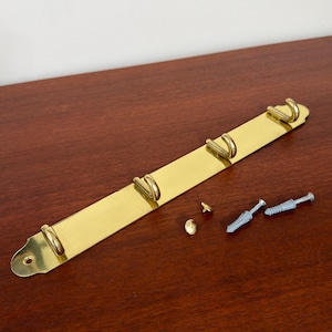 May include: A polished brass wall-mounted hook rack featuring four curved hooks attached to a long, flat rectangular plate with rounded ends. The set includes two small brass-colored screw covers and two grey plastic wall anchors for installation.
