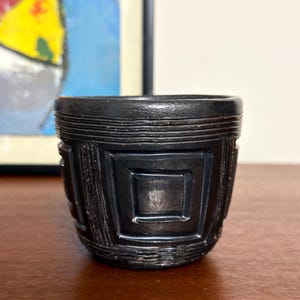 May include: A small, cylindrical black vessel featuring a textured surface with horizontal lines around the top and bottom rims. The exterior is decorated with a repeating geometric pattern of raised squares and vertical lines in a matte black finish.