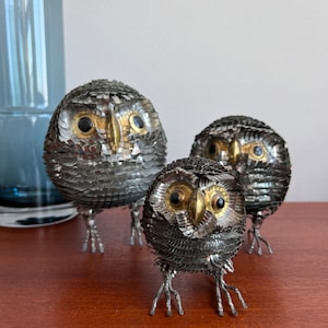 May include: Three silver metal owl sculptures with gold accents and black eyes. The owls are standing on a wooden surface.