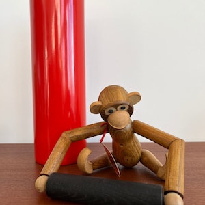 May include: A wooden monkey toy with movable arms and legs, holding a black rolling pin. The monkey has brown eyes and a red bow tie. The monkey is sitting on a wooden table next to a red cylinder.