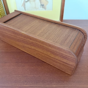 May include: A rectangular wooden box with a sliding lid. The box is made of light brown wood with visible grain patterns. The box is on a brown surface.