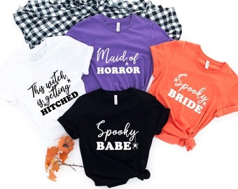May include: Four short-sleeve t-shirts in white, purple, black, and orange. The white shirt says "This witch is getting HITCHED", the purple says "Maid of HORROR", the black says "Spooky BABE", and the orange says "Spooky BRIDE" with spider icons.