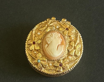 May include: A gold-coloured, round cameo pendant featuring a woman's profile. The cameo is set in an oval frame with floral and beaded details, and small red and turquoise accents. The pendant is set against a dark background.