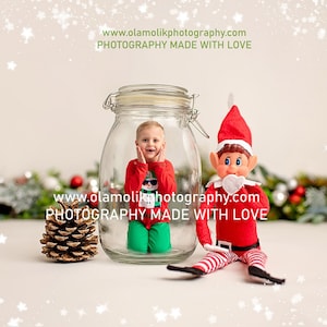 May include: A young child dressed in a red and green Christmas sweater is inside a clear glass jar. A Christmas elf doll is sitting next to the jar. The background is a white wall with a white sparkly overlay. The text "www.olamolikphotography.com PHOTOGRAPHY MADE WITH LOVE" is at the top of the image.