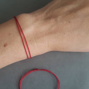 Könnte beinhalten: Nahaufnahme eines roten Armbands aus Faden, das am Handgelenk getragen wird, mit einem zweiten Armband darunter. Die Armbänder bestehen aus dünnem, leuchtend rotem Faden, und das Handgelenk ist hellhäutig.