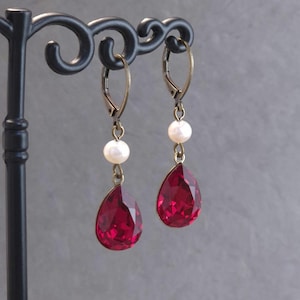 Peut inclure: Boucles d'oreilles pendantes avec des cristaux rouges en forme de goutte d'eau et des perles blanches. Les boucles d'oreilles ont des fermoirs à levier dorés. Les boucles d'oreilles sont exposées sur un support noir.