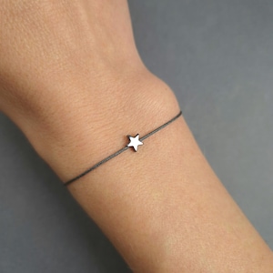 May include: A minimalist bracelet with a small, silver star charm. The star is strung on a thin, dark grey cord. The bracelet is displayed on a wrist, highlighting the simple design of the jewellery.