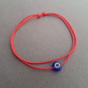 Könnte beinhalten: Ein rotes Armband mit einem blauen Glücksbringer in Form eines bösen Auges. Der Glücksbringer hat einen weißen Kreis in der Mitte.
