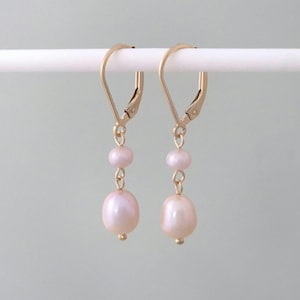 May include: A pair of gold dangle earrings with two pink pearls on each earring. The earrings have a leverback closure.