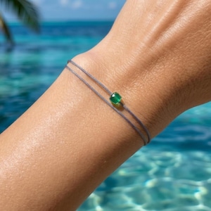 Könnte beinhalten: Ein zartes, zweireihiges Armband mit einer dünnen grauen Kordel und einer einzelnen kleinen, facettierten, quadratischen grünen Edelsteinperle, die mittig auf dem oberen Strang sitzt. Das Design betont den Kontrast zwischen der Kordel und dem Stein.