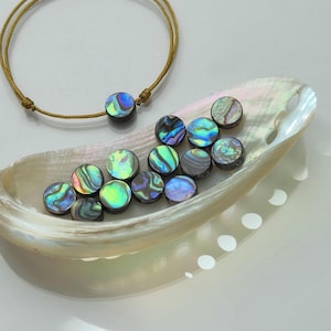 Peut inclure: Un bracelet en cordon doré avec une perle circulaire en nacre d'ormeau, accompagné d'une collection de perles d'ormeau circulaires et irisées disposées dans un grand coquillage d'ormeau incurvé et nacré.