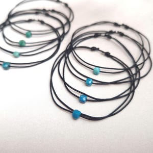 Könnte beinhalten: Mehrere schwarze Schnurarmbänder, jedes mit einer kleinen, türkisfarbenen Perle. Die Armbänder sind geknotet und scheinen handgefertigt zu sein. Die Perlen weisen eine Mischung aus Blau- und Grüntönen auf.