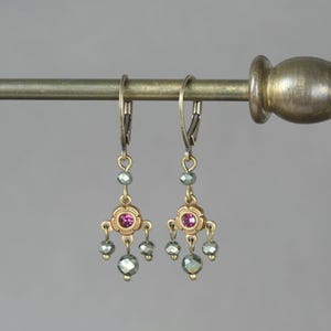 Peut inclure: Une paire de boucles d'oreilles pendantes avec une finition en laiton antique, chacune centrée par un petit cristal rose rond serti dans un cadre en forme de fleur, agrémentée de trois perles grises facettées suspendues en bas.