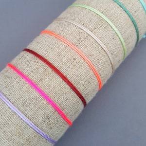 Peut inclure: Une collection de bracelets fins et colorés dans les tons rose, rouge, orange, vert et violet. Les bracelets sont présentés sur un tissu beige roulé, avec un fond gris. Les bracelets semblent être faits d'un matériau fin et tissé.
