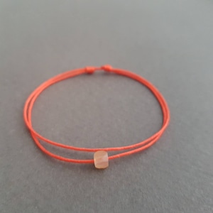 Könnte beinhalten: Ein minimalistisches Armband mit einer leuchtend orangefarbenen Kordel. In der Mitte der Kordel befindet sich eine kleine, hellfarbene, rechteckige Perle. Das Armband ist mit einem einfachen Knotenverschluss versehen. Der Hintergrund ist einfarbig grau.