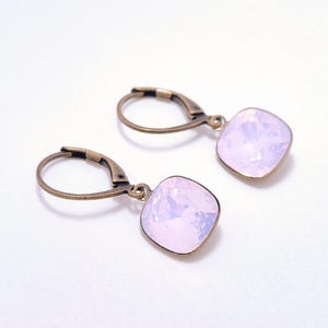 Peut inclure: Une paire de boucles d'oreilles avec des pendentifs en cristal rose clair de forme carrée. Les boucles d'oreilles ont des fermoirs à levier de couleur bronze. Les cristaux ont une qualité irisée, reflétant la lumière. Sur fond blanc.