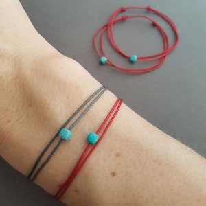 Könnte beinhalten: Zwei dünne Kordelarmbänder, eines in Dunkelgrau und eines in Rot, jeweils mit einer kleinen, quadratischen, türkisfarbenen Perle. Die Armbänder werden am Handgelenk getragen, während zwei weitere rote Kordelarmbänder mit türkisfarbenen Perlen daneben liegen.