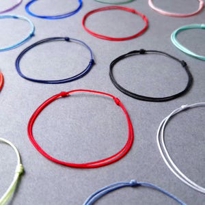 Op de afbeelding: Een assortiment kleurrijke touwarmbanden in verschillende kleuren, waaronder rood, blauw, zwart en wit, liggen op een grijze ondergrond. Elke armband heeft een eenvoudig, verstelbaar knoopontwerp, wat een minimalistische esthetiek laat zien.