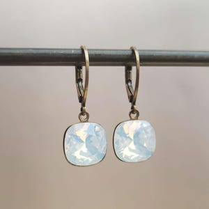 Peut inclure: Une paire de boucles d'oreilles carrées avec une pierre irisée bleu clair sertie dans un métal de couleur bronze. Les boucles d'oreilles sont suspendues à une fermeture à levier. Les boucles d'oreilles sont présentées sur une barre métallique.