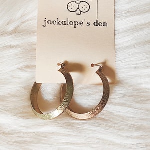 May include: A pair of gold-colored hoop earrings with etched floral patterns. The earrings are displayed on a tag with the text "jackalope's den" and a whimsical illustration of antlers, a monocle, and a rabbit face.