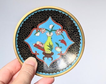 May include: Circular decorative coaster with a gold edge. The coaster has a central design of a light blue shape with a green vase holding flowers, set against a black patterned background. The coaster is held by a hand.