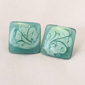 Puede incluir: Un par de pendientes cuadrados. Cada pendiente es de color verde azulado claro con un diseño blanco nacarado en espiral. Los pendientes tienen una forma ligeramente redondeada y un acabado brillante. Los pendientes se muestran sobre una superficie blanca.