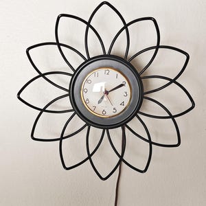 May include: A black metal flower-shaped wall clock with a round clock face. The clock face has a white background with black numbers and hands. The clock is hanging on a white wall.