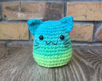 May include: A handmade crochet cat in shades of blue and green. The cat has black button eyes and a stitched mouth and whiskers. The cat is sitting on a wooden surface with a brick background.