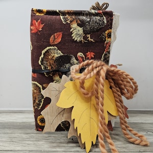 May include: A handmade journal with a brown turkey and fall leaves pattern. The journal is tied with a black elastic band and adorned with wooden leaf cutouts and a thick, tan rope. The journal's cover features a repeating pattern of turkeys, sunflowers, and autumn leaves.