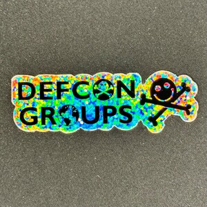 Op de afbeelding: Een holografische sticker met de tekst "DEFCON GROUPS" en een schedel met gekruiste beenderen met een smiley.
